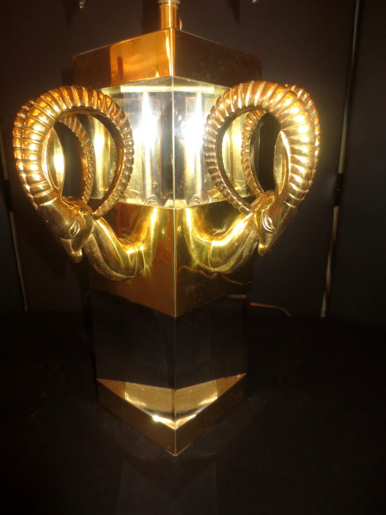 May include: A gold-colored table lamp with a geometric design. The lamp features two ram's head sculptures on the sides. The lamp has a rectangular base and a gold and black color scheme.