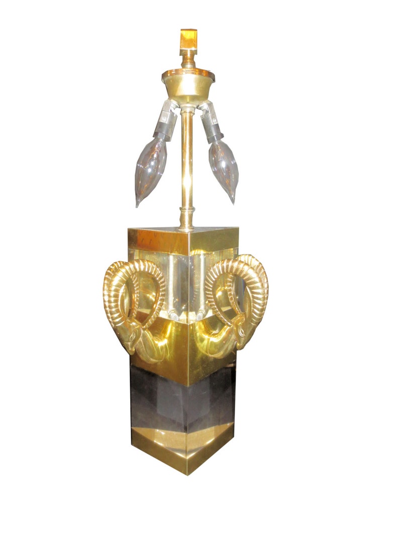 May include: A vintage brass table lamp with a geometric design. The lamp features a square base with gold accents and a black center section. Two decorative ram's horn shapes adorn the front. The lamp has three elongated light bulbs.