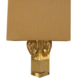 May include: A gold-toned lamp with a rectangular lampshade in a tan color. The lamp base features a geometric design with decorative ram's head accents. The lamp is designed for indoor use and has a classic, elegant style.