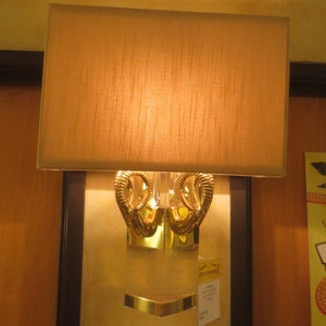 May include: A wall-mounted lamp with a rectangular, tan lampshade. The lamp features a gold-colored, ornate base with a decorative design. A small, gold-colored shelf is positioned below the base. The lamp is mounted on a light-colored wall.