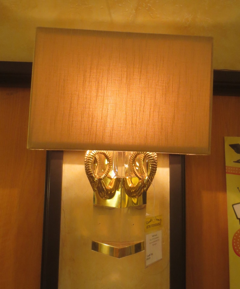 May include: A wall-mounted lamp with a rectangular, tan lampshade. The lamp features a gold-colored, ornate base with a decorative design. A small, gold-colored shelf is positioned below the base. The lamp is mounted on a light-colored wall.
