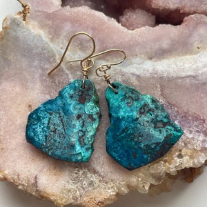 May include: A pair of turquoise green earrings with gold wire accents. The earrings are made of natural stone and have a unique, organic shape.