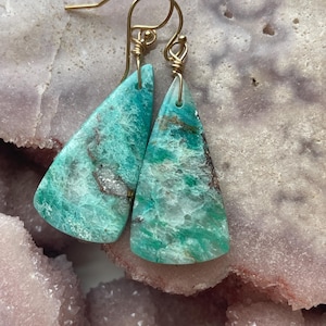 May include: A pair of turquoise gemstone earrings with gold-colored ear wires. The earrings are triangular, with a marbled pattern of green, brown, and white. The earrings are displayed on a light purple surface.