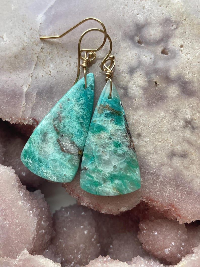 May include: A pair of turquoise gemstone earrings with gold-colored ear wires. The earrings are triangular, with a marbled pattern of green, brown, and white. The earrings are displayed on a light purple surface.