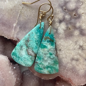 May include: A pair of handcrafted earrings featuring triangular, turquoise-colored stones with gold-colored ear wires. The stones have a marbled appearance with shades of green and brown, set against a light pink and gray background.