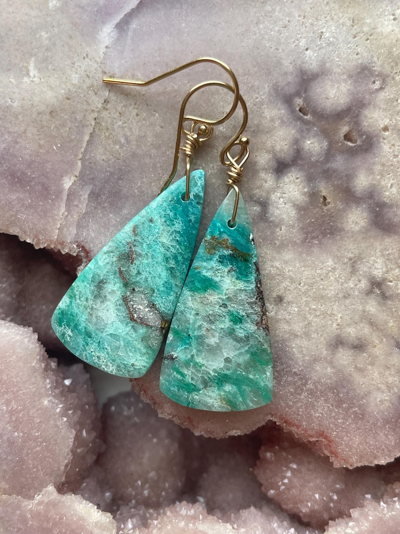 May include: A pair of handcrafted earrings featuring triangular, turquoise-colored stones with gold-colored ear wires. The stones have a marbled appearance with shades of green and brown, set against a light pink and gray background.