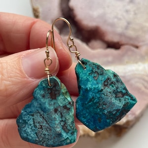 May include: A pair of turquoise green gemstone earrings with gold wire wrapped around the stones and gold hooks. The stones have a rough, natural texture.