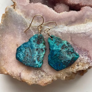 May include: A pair of turquoise blue gemstone earrings with gold wire wrapped around the stones and gold hooks. The earrings are displayed on a pink and white crystal background.