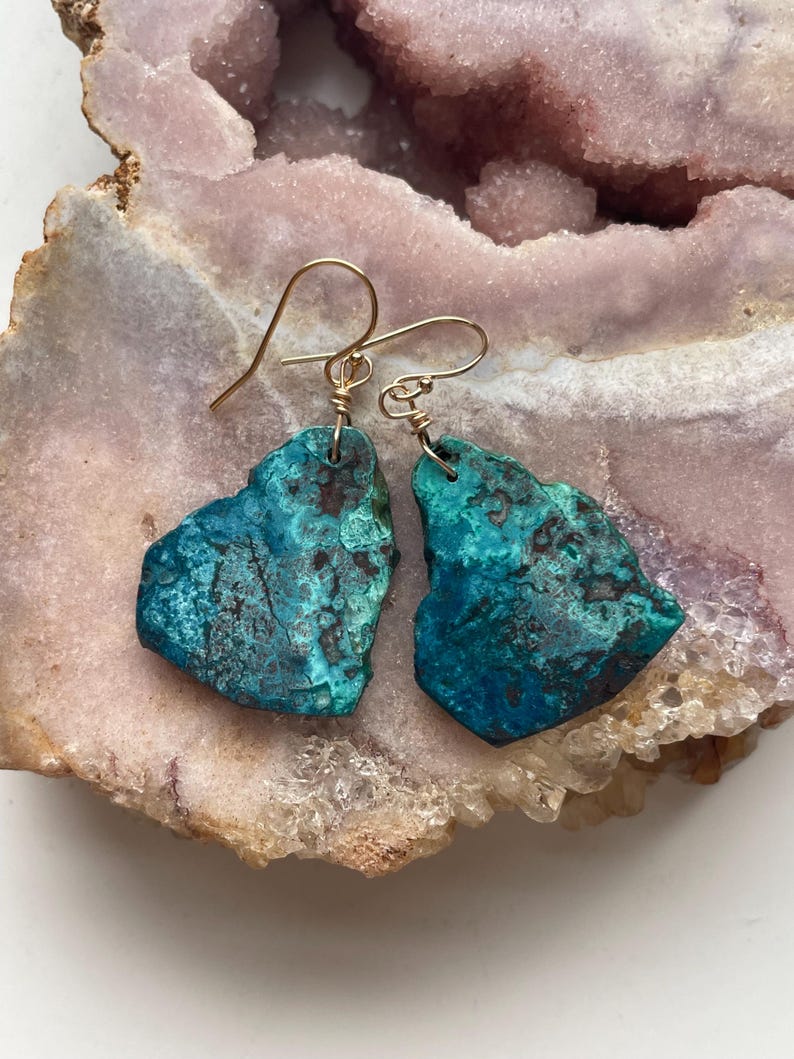 May include: A pair of turquoise blue gemstone earrings with gold wire wrapped around the stones and gold hooks. The earrings are displayed on a pink and white crystal background.