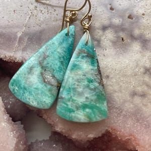 May include: A pair of earrings featuring triangular, turquoise-colored stones with gold-colored wire hooks. The stones have a marbled appearance with shades of green, brown, and white. The earrings are displayed on a pink and white surface.
