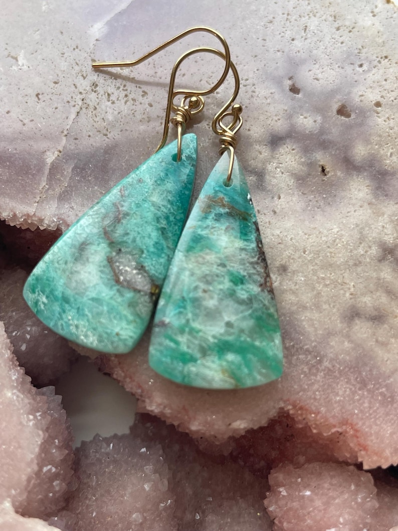 May include: A pair of earrings featuring triangular, turquoise-colored stones with gold-colored wire hooks. The stones have a marbled appearance with shades of green, brown, and white. The earrings are displayed on a pink and white surface.