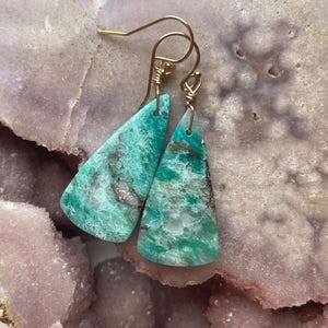 May include: A pair of turquoise gemstone earrings with gold-colored ear wires. The earrings are triangular, with a mottled pattern of green, brown, and white. The earrings are displayed on a light purple and pink surface.