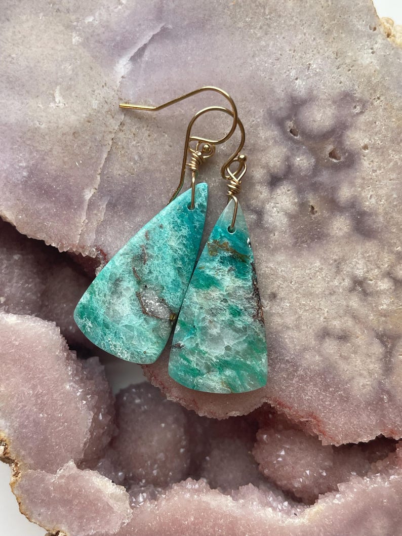 May include: A pair of turquoise gemstone earrings with gold-colored ear wires. The earrings are triangular, with a mottled pattern of green, brown, and white. The earrings are displayed on a light purple and pink surface.