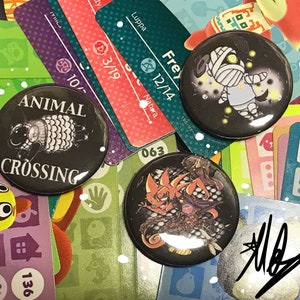 Peut inclure: Trois boutons sur le thème d'Animal Crossing avec des fonds noirs. Le premier bouton représente un mouton noir et blanc avec le texte "ANIMAL CROSSING". Le deuxième bouton représente une momie blanche sur un fond noir avec des étoiles jaunes. Le troisième bouton représente une créature rouge et noire sur un fond noir.