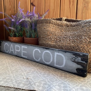 May include: A wooden sign with the words "CAPE COD" painted in white on a grey background. The sign is distressed and has a black border. The sign is sitting on a white rug in front of a wooden wall.