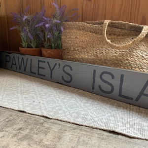 Può includere: Un cartello in legno grigio con il testo "PAWLEY'S ISLAND" in lettere nere. Il cartello è appoggiato su un tappeto bianco davanti a un cesto intrecciato e piante in vaso.