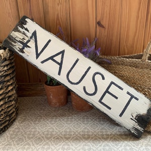 Könnte beinhalten: Ein weißes Holzschild mit schwarzer Beschriftung, auf dem "NAUSET" steht. Das Schild ist abgenutzt und hat einen rustikalen Look. Es lehnt an einem geflochtenen Korb.