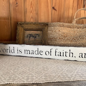 Puede incluir: Un letrero de madera blanco con letras negras que dice "The world is made of faith, and ..." El letrero está sobre una alfombra blanca frente a una pared de madera. Hay una pequeña imagen enmarcada de un caballo y una cesta tejida en el fondo.