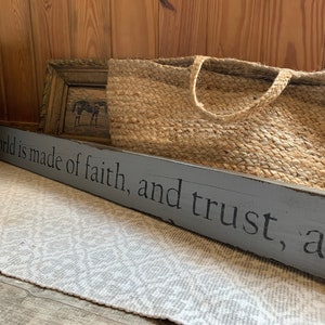 Puede incluir: Un letrero de madera gris con el texto "The world is made of faith, and trust, and..." El letrero está sobre una alfombra blanca con una cesta tejida y un marco de imagen marrón en el fondo.