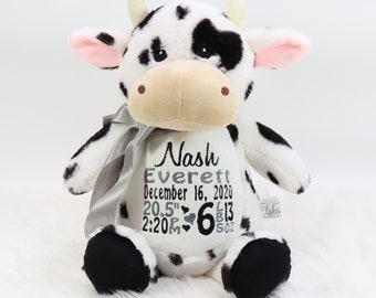 May include: A plush cow with black spots, a beige snout, and pink inner ears. The cow has a silver ribbon and embroidered text that reads "Nash Everett December 16, 2020 20.5" 6 L13 OZ 2:20P".