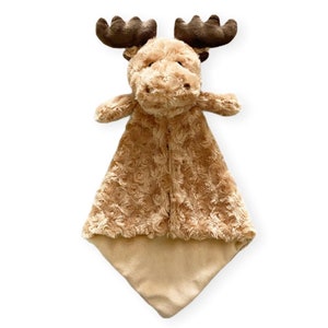 May include: A plush, tan-colored moose security blanket. The moose has brown antlers, a soft face, and small arms. The blanket portion is a textured, rose-patterned fabric that transitions to a smooth, tan square at the bottom. A comforting item for babies and toddlers.