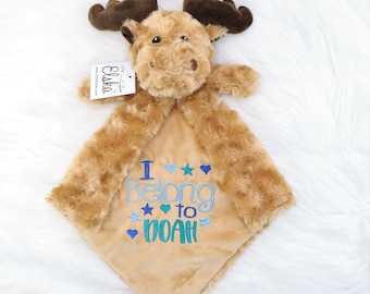May include: A plush moose-shaped baby blanket in shades of brown and tan. The blanket features embroidered text that reads "I Belong to NOAH" in blue and teal. The moose has brown antlers and a tag that reads "Elska".