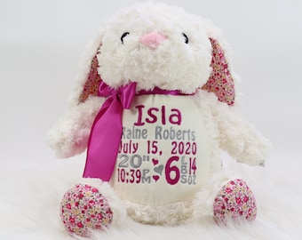 May include: A white plush bunny with pink accents, including a bow and floral patterned ears and feet. The bunny has embroidered details and a personalized panel with the name "Isla", birthdate, and measurements. The bunny is approximately 20 inches tall.