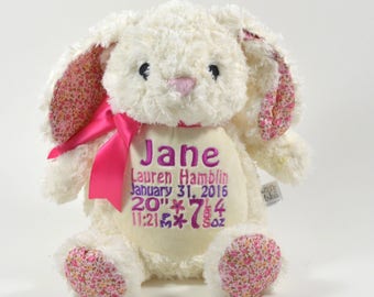 May include: A plush white bunny rabbit with pink floral patterned ears, feet, and accents. The bunny has a pink bow and embroidered text that reads "Jane" and other details. The bunny is approximately 20 inches tall.