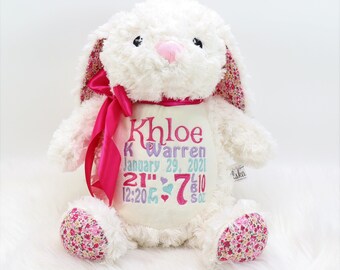 May include: A white plush bunny with a pink bow and floral accents on the ears and feet. The bunny has embroidered details, including a name, date, and measurements. The bunny is 21 inches tall and weighs 7 lbs 10 oz.