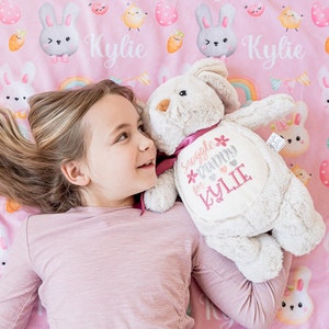 May include: A soft, light grey bunny toy with a pink ribbon and embroidered text reading "Snuggle Bunny for Kylie." The bunny rests on a pink blanket decorated with bunny and rainbow patterns, and the name "Kylie".