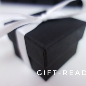 May include: A black gift box with a white ribbon tied around it. The box is sitting on a white surface. The text "GIFT-READY" is printed on the surface below the box.