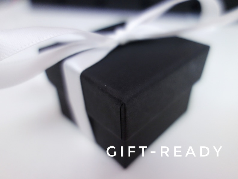 May include: A black gift box with a white ribbon tied around it. The box is sitting on a white surface. The text "GIFT-READY" is printed on the surface below the box.