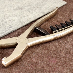 May include: A silver metal punch tool with a black handle and multiple interchangeable punch heads. The tool is lying on a brown surface.