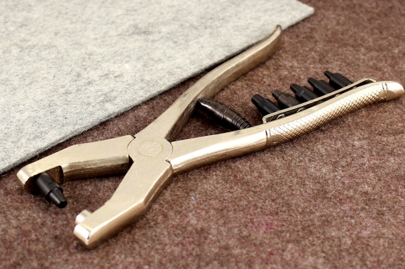 May include: A silver metal punch tool with a black handle and multiple interchangeable punch heads. The tool is lying on a brown surface.