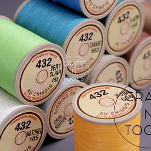 May include: Close-up of spools of thread in various colors, including green, blue, yellow, and white. The spools are labeled with the number "432" and the text "Au Chinois" in French. The spools are arranged in a stack, with the top spool being green.