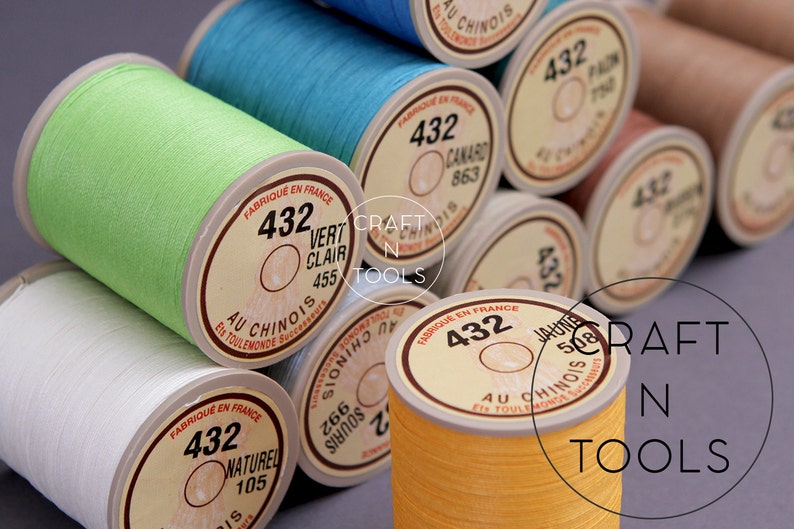 May include: Close-up of spools of thread in various colors, including green, blue, yellow, and white. The spools are labeled with the number "432" and the text "Au Chinois" in French. The spools are arranged in a stack, with the top spool being green.