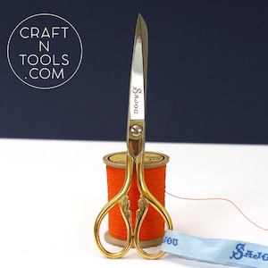May include: Gold-coloured embroidery scissors with a decorative handle and the word "Sasou" engraved on the blade. The scissors are resting on a spool of orange thread.
