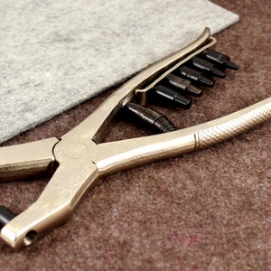 May include: A silver metal punch tool with a black handle and a set of interchangeable punches. The tool is used for making holes in leather, fabric, or other materials.