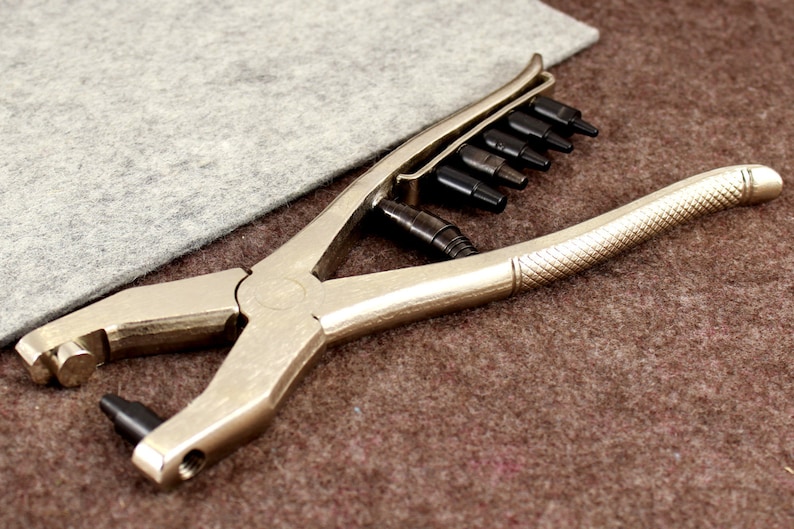 May include: A silver metal punch tool with a black handle and a set of interchangeable punches. The tool is used for making holes in leather, fabric, or other materials.