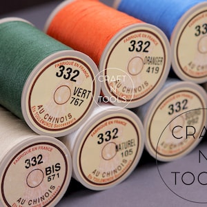 Peut inclure: Gros plan sur des bobines de fil de différentes couleurs, notamment vert, orange, bleu, beige et blanc. Chaque bobine porte une étiquette avec le texte "332" et un nom de couleur en français. L'étiquette comprend également le texte "Au Chinois" et "Fabrique en France".