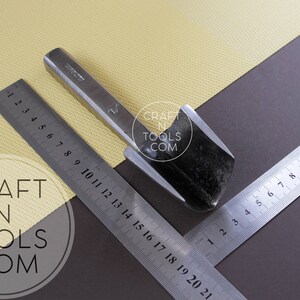 Puede incluir: Un primer plano de una herramienta de trabajo del cuero de metal con una cuchilla curva. La herramienta est&aacute; sobre una superficie amarilla con una regla marcada en cent&iacute;metros. El texto "CRAFT N TOOLS COM" es visible en la imagen.