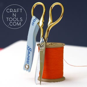 May include: Gold-coloured embroidery scissors with a blue ribbon and a spool of orange thread.