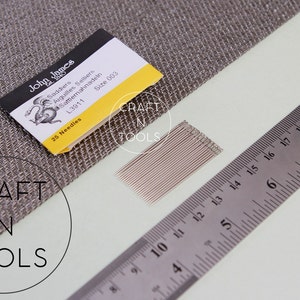 May include: A pack of 25 John James Saddlers needles, size 003, with a yellow and black label. The needles are laid out on a white surface next to a ruler.