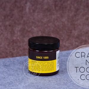 May include: A small, round jar with a black lid and a yellow label. The label says "Since 1895" and has instructions for using the product. The jar is sitting on a gray surface.
