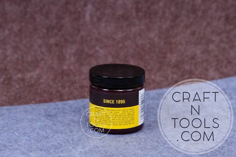 May include: A small, round jar with a black lid and a yellow label. The label says "Since 1895" and has instructions for using the product. The jar is sitting on a gray surface.
