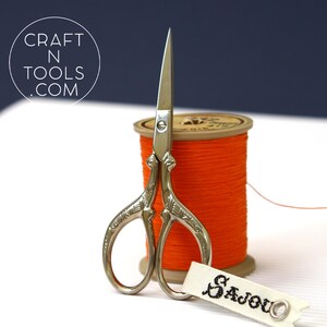 May include: Silver embroidery scissors with ornate handles, next to a spool of orange thread and a white label with the text "Sajou" in black.