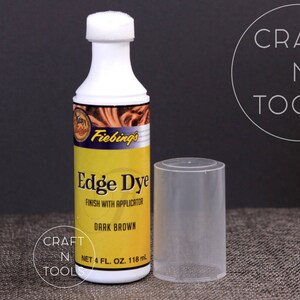 以下が含まれることがあります： Fiebing's Edge Dye ダークブラウンのボトル。革の縁を仕上げるための染料です。ボトルは118mlで、アプリケーターが付いています。ボトルは茶色の表面に置かれ、隣には透明なプラスチックチューブがあります。