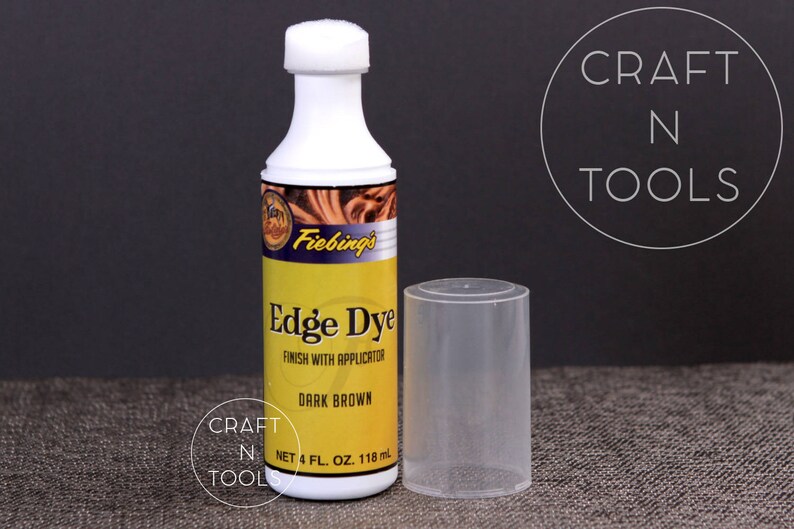 以下が含まれることがあります： Fiebing's Edge Dye ダークブラウンのボトル。革の縁を仕上げるための染料です。ボトルは118mlで、アプリケーターが付いています。ボトルは茶色の表面に置かれ、隣には透明なプラスチックチューブがあります。