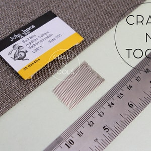 May include: A pack of 25 silver saddler needles, size 005, from John James, labeled "Saddlers Aiguilles Selliers Sattlernahnadein L3911 Size 005 25 Needles". The needles are arranged in a row and are next to a ruler and a piece of fabric.