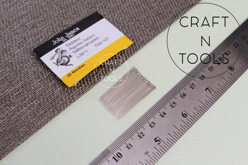 May include: A pack of 25 silver saddler needles, size 005, from John James, labeled "Saddlers Aiguilles Selliers Sattlernahnadein L3911 Size 005 25 Needles". The needles are arranged in a row and are next to a ruler and a piece of fabric.